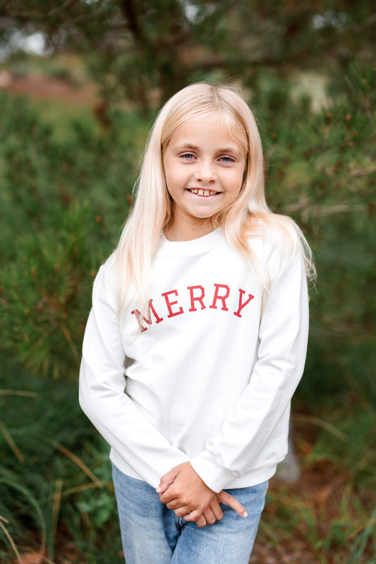Young girl wearing a white sweatshirt with 'MERRY' in red text, standing outdoors with greenery in the background.