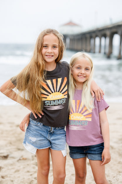 Two young girls standing on a beach wearing matching t-shirts with sun designs and 'Cruel Summer' text.