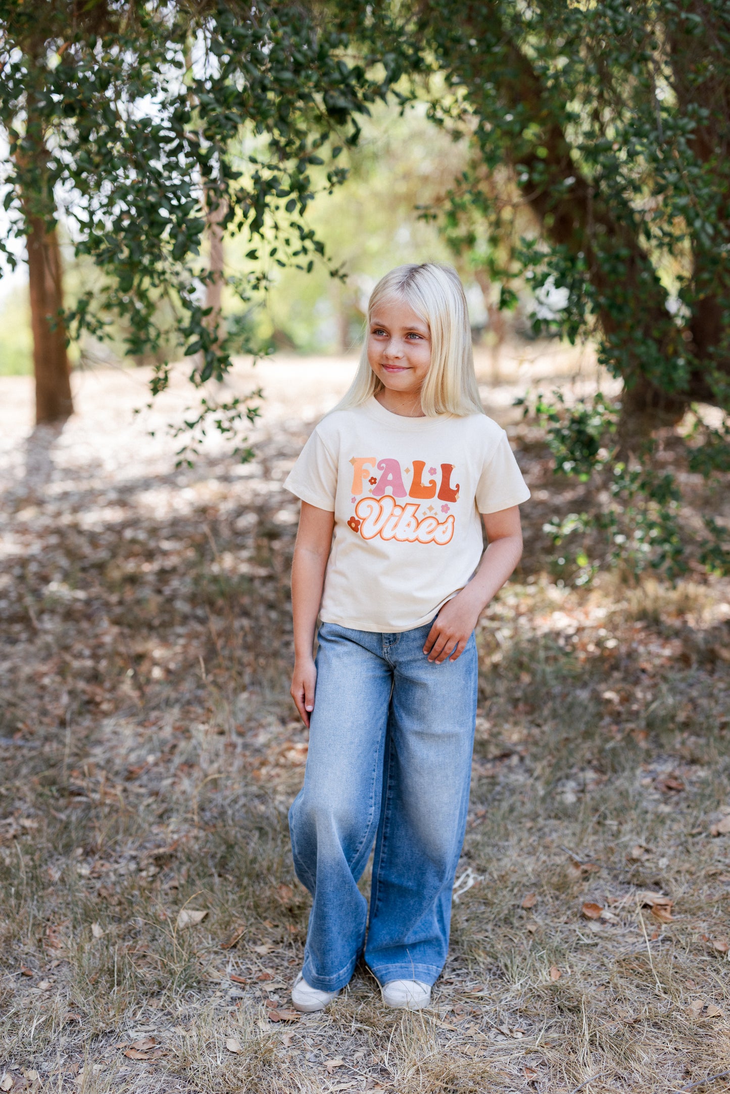Young girl wearing a 'Fall Vibes' t-shirt standing in a natural setting with trees and grass.