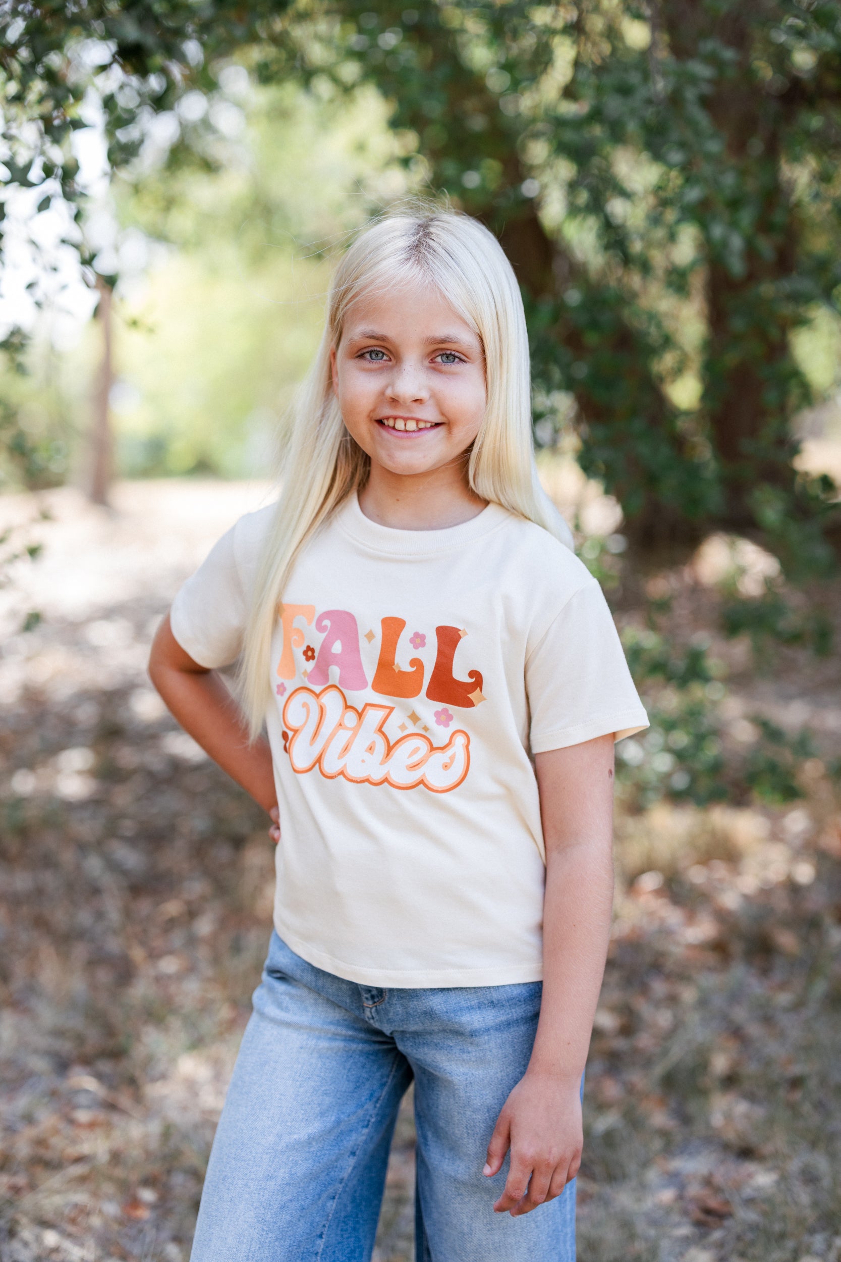 Young girl wearing a 'Fall Vibes' t-shirt outdoors with trees in the background