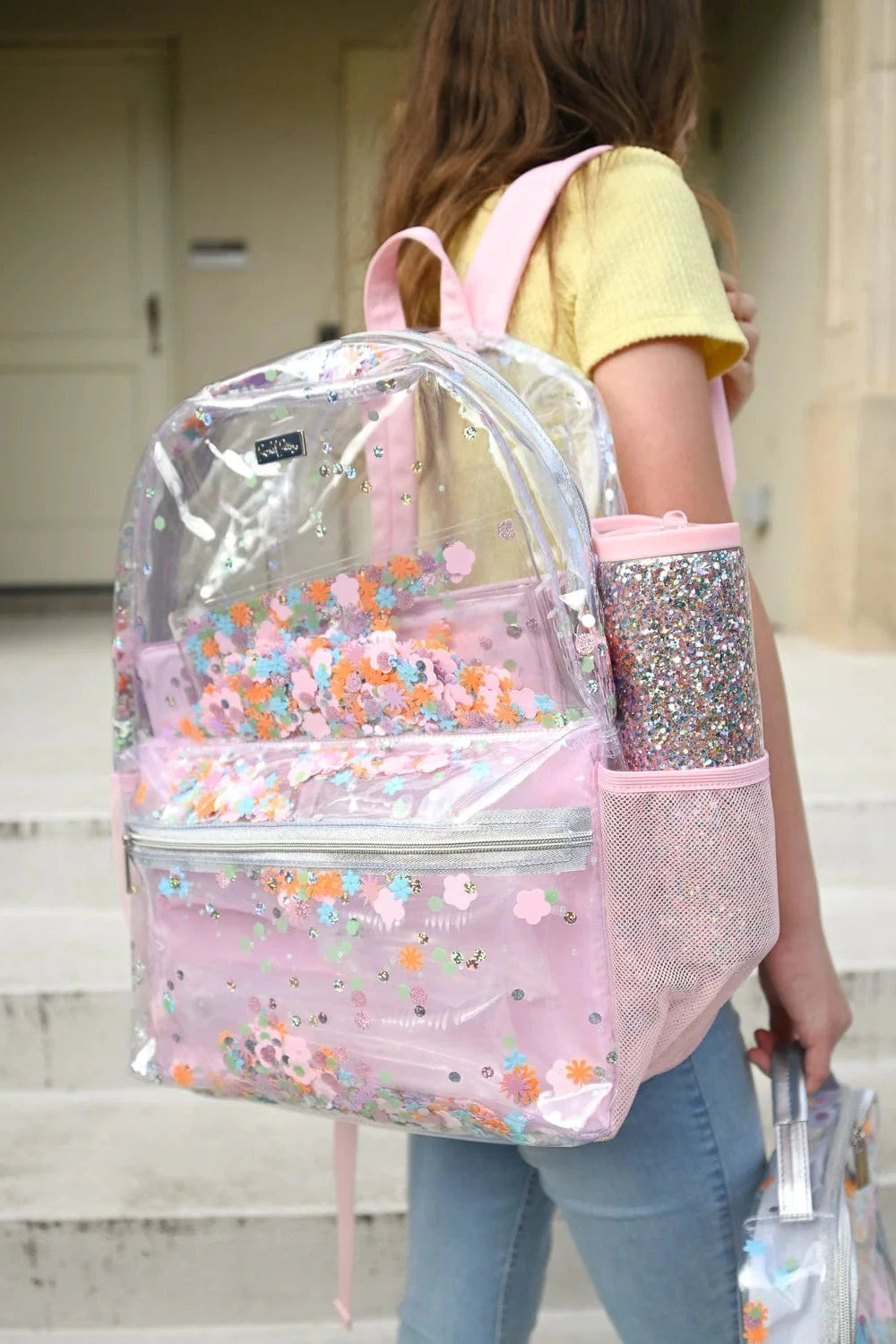 Person wearing a clear backpack with pink accents and floral designs, holding a glittery water bottle.