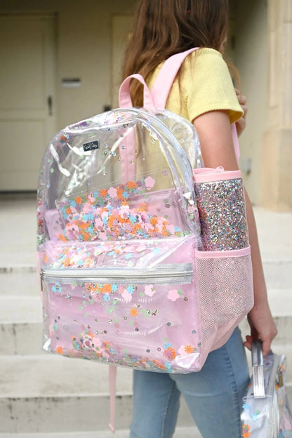 Person wearing a clear backpack with pink accents and floral designs, holding a glittery water bottle.