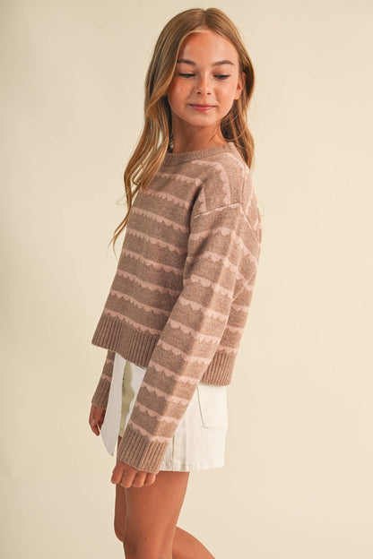 A girl wearing a cropped, long-sleeve knit sweater with horizontal wavy stripes in shades of taupe and light brown