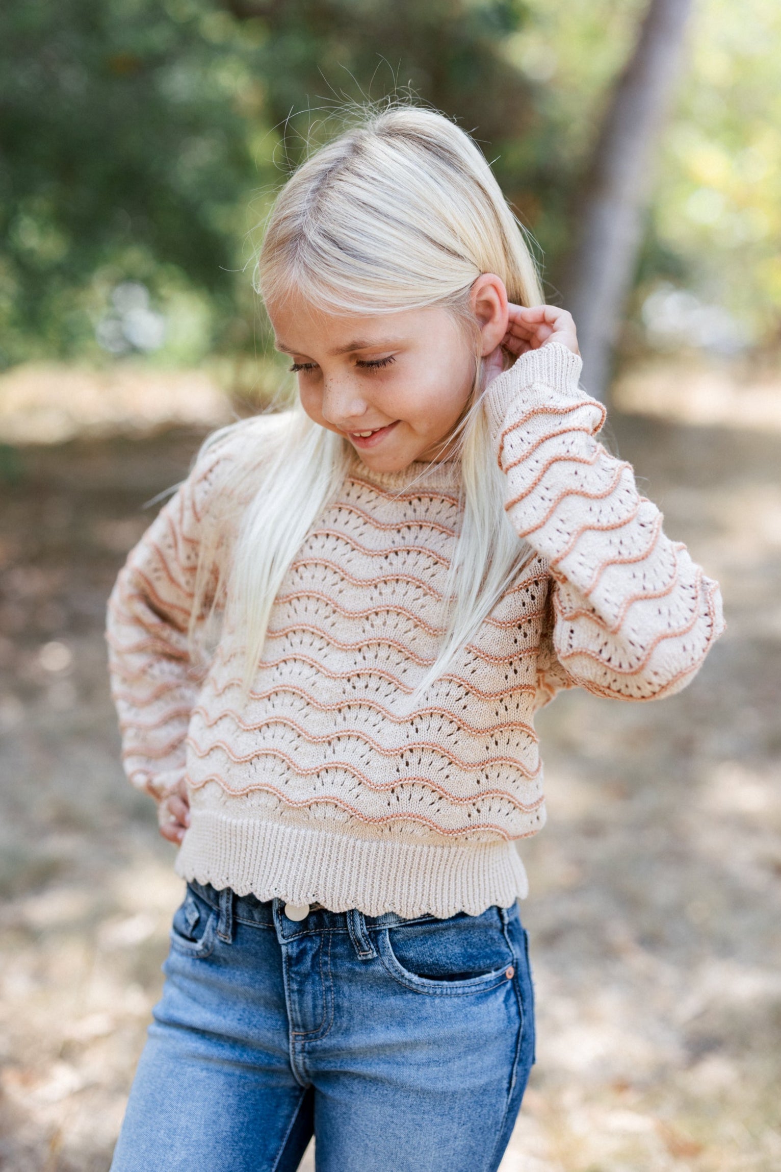 Young girl wearing a beige knitted sweater and blue jeans outdoors.
