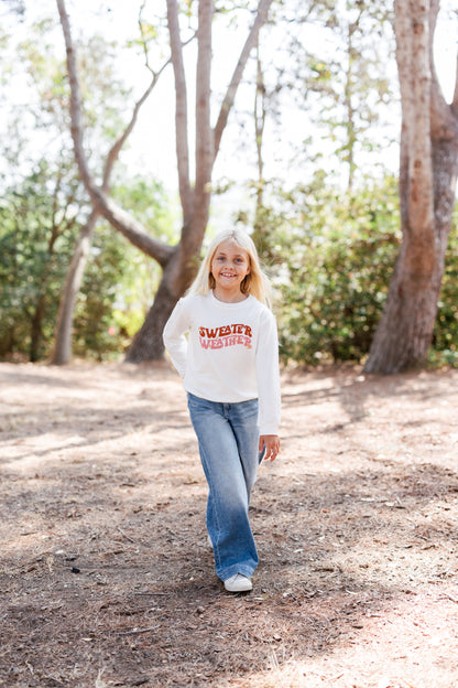 Girl walking outdoors in a wooded area wearing a white sweatshirt with text "SWEATER WEATHER"