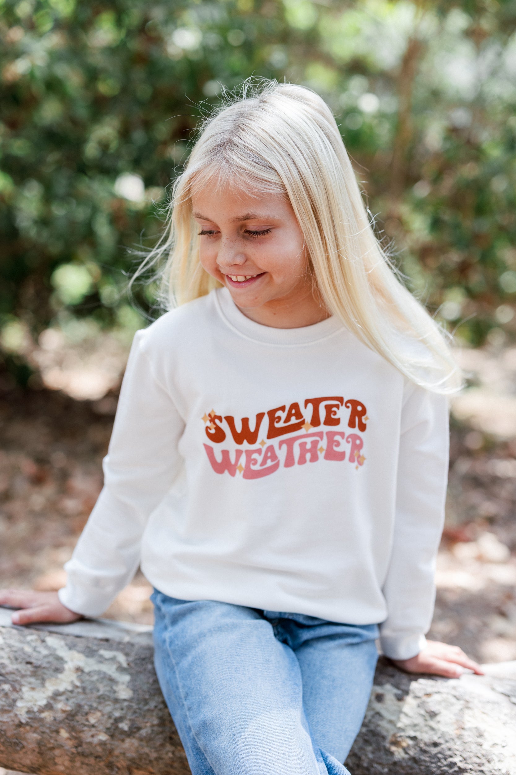 Girl wearing a white sweatshirt with 'Sweater Weather' text, sitting outdoors.