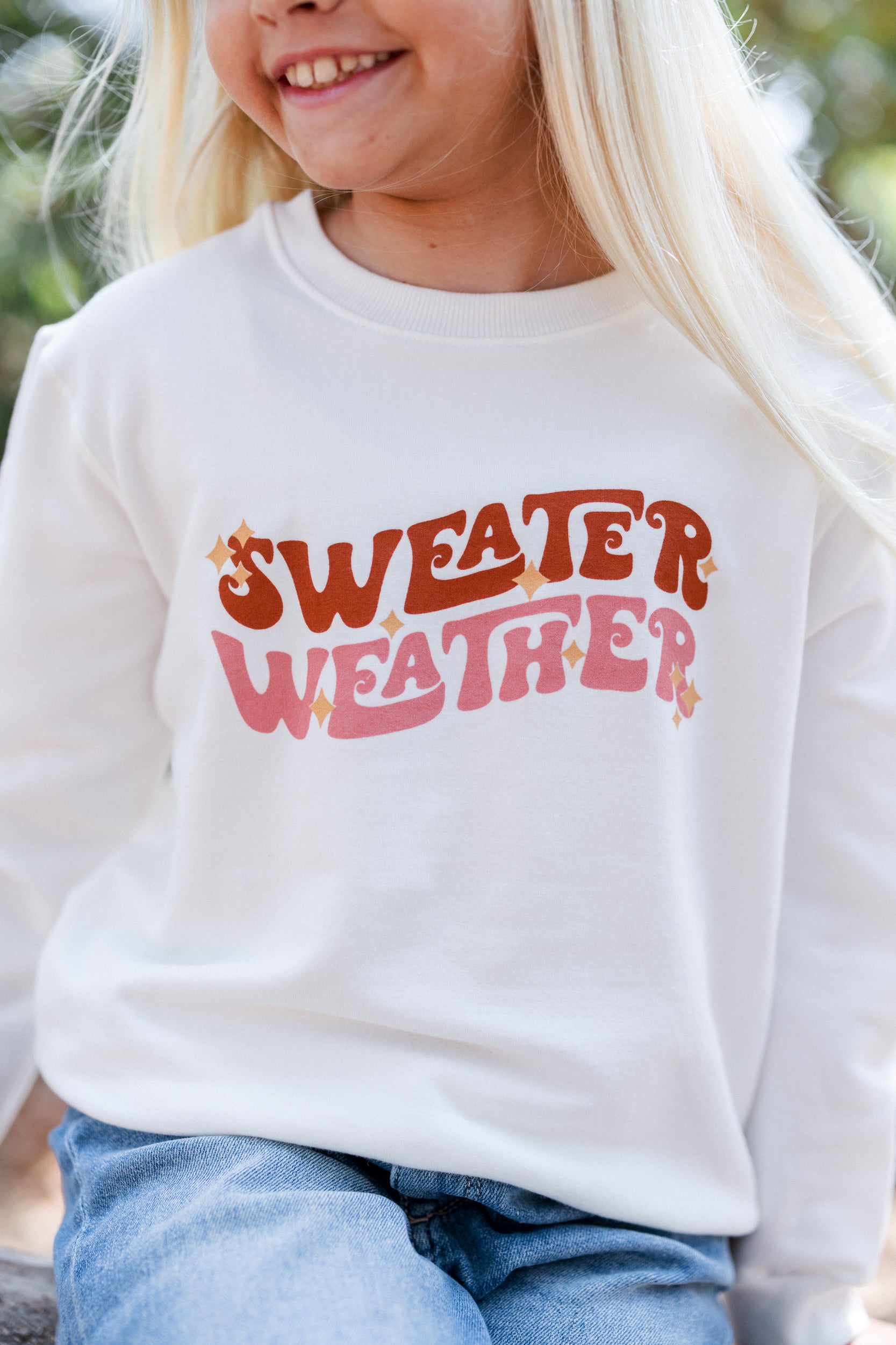 Girl wearing a white sweatshirt with 'SWEATER WEATHER' text, sitting outdoors.